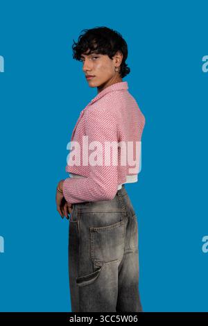 Fashionable young latin model looking over shoulder while wearing pink cropped jacket and grey jeans against blue background Stock Photo