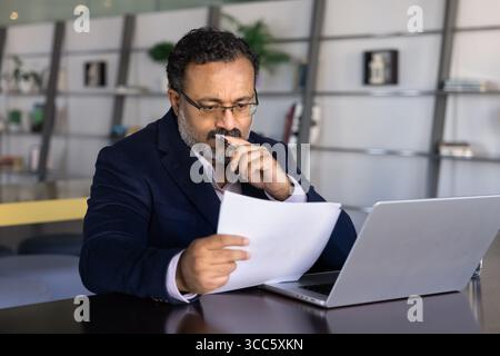 Thoughtful frowning senior Indian businessman reading paper document at laptop Stock Photo