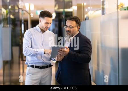 Serious younger software professional giving help to senior Indian colleague Stock Photo