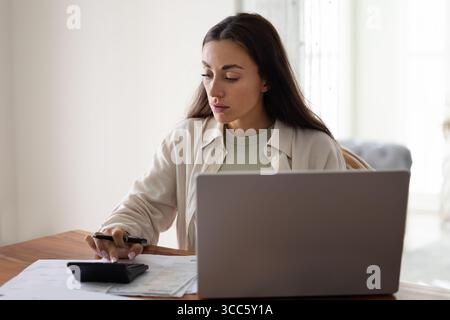 Focused young woman sits at table using calculator and laptop Stock Photo