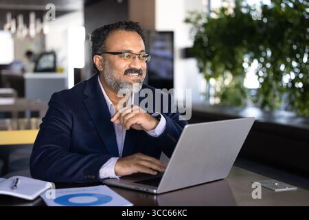 Positive successful mature Indian executive man looking away from workplace Stock Photo