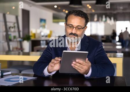 Serious elder Indian executive man typing on tablet at table Stock Photo