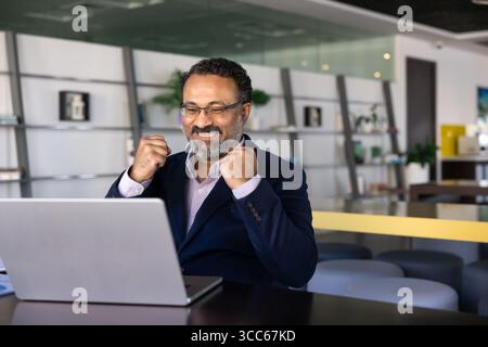 Joyful senior Indian product owner making winner fists at computer Stock Photo