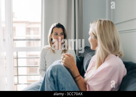 Two cheerful beautiful blonde women are chatting while sitting on the sofa in the living room. Stock Photo