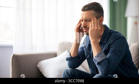 Stressed man having headache, sitting on sofa at home Stock Photo