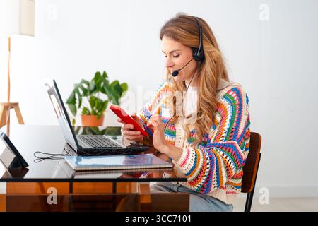Woman with headset working on laptop and using smartphone at home office Stock Photo