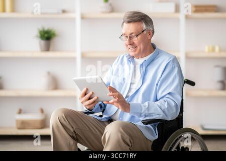 Joyful impaired senior man in wheelchair using tablet computer at home Stock Photo
