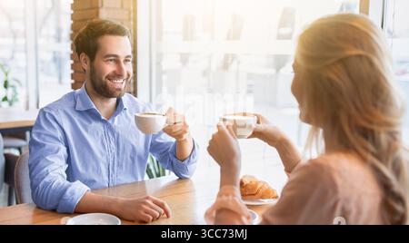 Cheerful young guy having coffee together with his girlfriend at cafe, chatting and smiling during breakfast Stock Photo