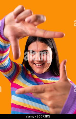 Cheerful young woman playfully framing her face with hands and smiling against a vibrant orange backdrop, showcasing creativity and positivity in a co Stock Photo