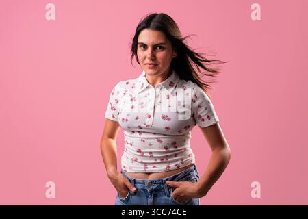 Young woman with unique style posing in a professional studio, hair blowing in the wind from a fan, against a vibrant pink backdrop and bright studio Stock Photo