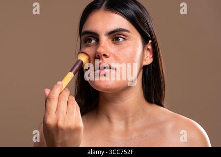 Concentrated young woman with freckles is applying makeup on her face using a brush, promoting skincare and body care routines for natural beauty enha Stock Photo