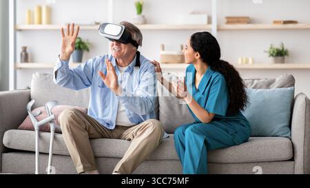Disabled senior man using VR headset with young nurse, exploring augmented reality at home Stock Photo