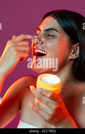 Radiant woman joyfully applying skincare cream to her nose, holding a jar against a vibrant background. Emphasizing beauty, self-care, and healthy ski Stock Photo