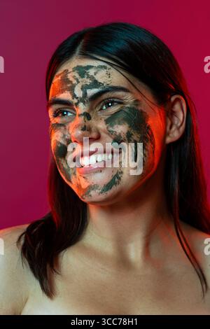 Studio portrait of a cheerful young woman with dark hair undergoing a beauty treatment, wearing a green mud facial mask and smiling, promoting skin ca Stock Photo