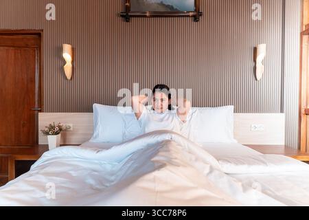 A young Indonesian woman sitting on bed in the morning, wearing a white t-shirt and smiling. Suitable for lifestyle, morning routine, bedroom, Stock Photo