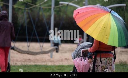 Wroclaw, Poland, August 2025 Woman holding a rainbow colored umbrella and pushing a stroller on a playground during rainy weather, with children playi Stock Photo