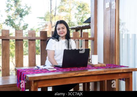 An Indonesian woman sitting at a wooden table on an outdoor terrace, working on her laptop with a relaxed smile. Stock Photo