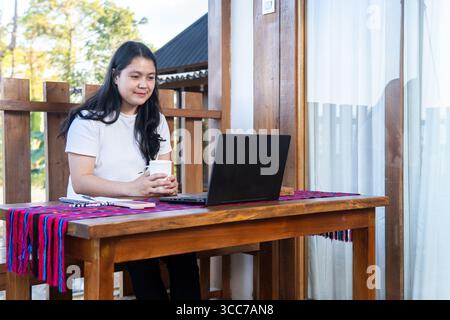 A young Asian woman in a white t-shirt works on a laptop at a wooden desk outdoors, writing notes and smiling. Remote work, freelance lifestyle. Stock Photo