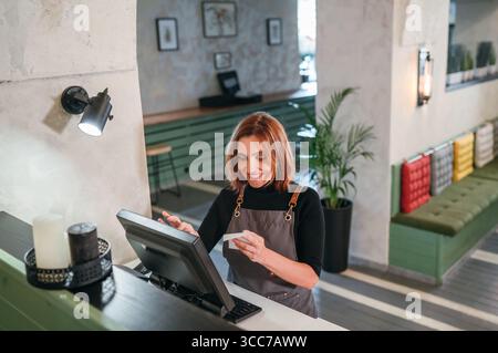 Smiling waitress in black uniform using point-of-sale terminal to process customer orders. Bartender at work in restaurant, hospitality service, and g Stock Photo