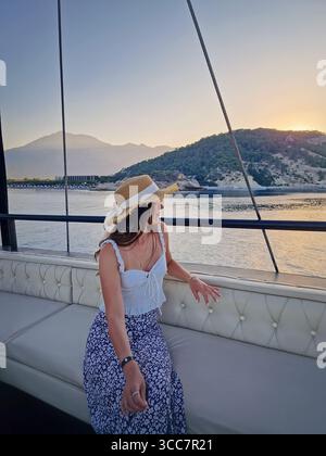 Young woman tourist wearing straw hat, sailing on a boat gazing out the Kiris resort coastline in Turkey. Female enjoying the views as cruising on the Stock Photo
