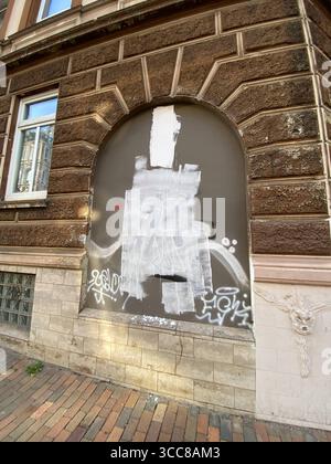 Flensburg, Schleswig-Holstein Vergraffitiertes, zugemauertes Rundbogenfenster an einer historischen Fassade in Flensburg, Friesische Straße. Diverse tags und weiße Farbflecken überdecken die ehemals glatte Oberfläche. Das Übermalte zeigte zuvor eine stehende Figur in einem grob mit schwarzer Farbe gemalten Sträflingsanzug mit vertikalen Streifen. Die Person trug einen angedeuteten Turban und hielt vor der Brust ein großes Stück Wassermelone, dessen leuchtende Farben grün, rot, schwarz im Kontrast zum schwarz-weißen Restbild. Die Haltung wirkte verschlossen, da die Arme vor dem Oberkörper versc Stock Photo