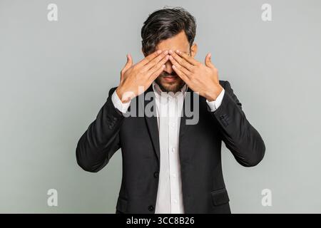 Don't want to look at this, awful. Afraid shocked Indian businessman closing eyes with hand showing stop gesture, confused shy scared reject to watch. Male Arabian man guy isolated on gray background Stock Photo