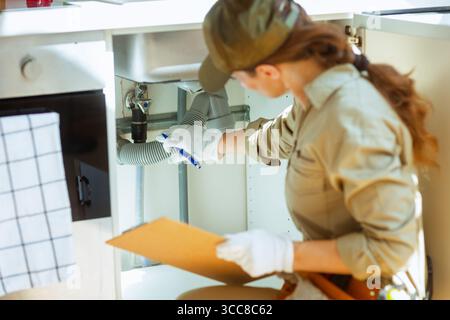A focused plumber in a tan uniform inspects under a kitchen sink, taking notes on a clipboard during a service call or home inspection. Stock Photo