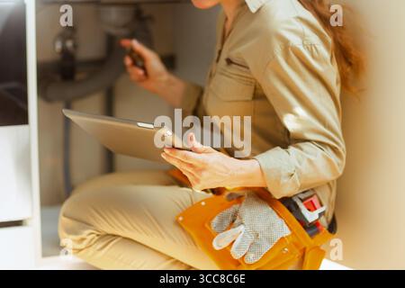 A woman in tan work clothes kneels under a sink, using a tablet to inspect plumbing parts. She's likely diagnosing a leak or other problem. Stock Photo