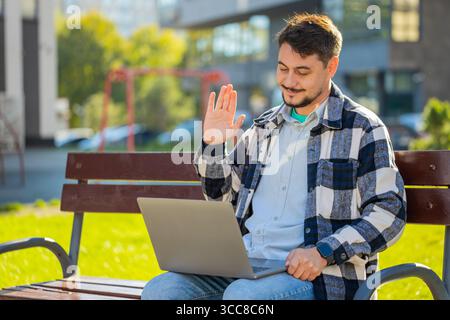 Mature businessman talking on video call using laptop computer while sitting on bench in city street. Male manager employer in casual clothes having an online conversation, remote business meeting. Stock Photo