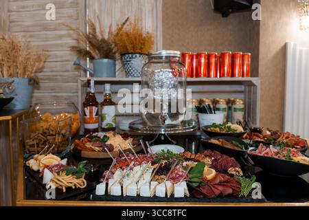 A lavish charcuterie board assortment with cubed cheese, cured meats, olives, dips, and canned beverages arranged on a dark wood table for an event Stock Photo