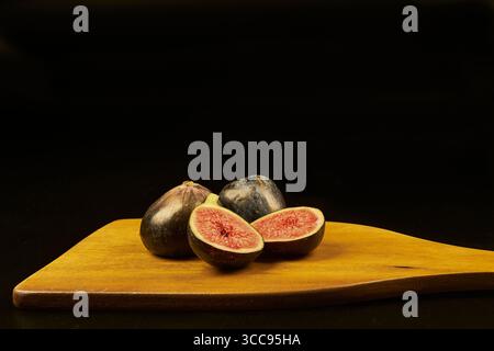 Fresh figs on a black background arranged on a wooden board, some cut in half showing the beautiful red color of their flesh. Stock Photo