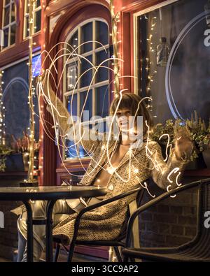 Beautiful young woman sitting in front of building decorated with fairy lights, while doing light painting effect by moving and turning lights in hand Stock Photo