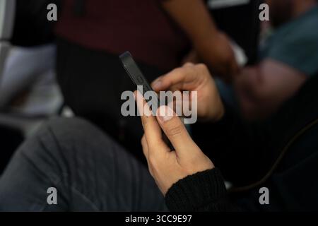 Close up of a Latino male's hands using a smartphone, while seated on an airplane as people board in the background. Stock Photo