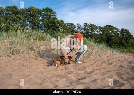 Engaged pet owner playing with cat on riverbank sand first outdoor walk. Animal curiosity and fear Stock Photo
