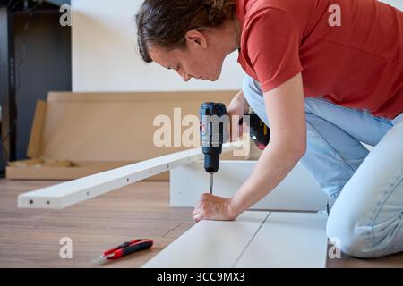 Woman using cordless drill to screw part into white bed frame. Female assembling furniture in new apartment. DIY home renovation and home organization Stock Photo