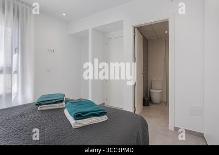 Minimalist bedroom featuring a neatly made bed with folded towels, direct access to a modern ensuite bathroom, and bright natural light through sheer Stock Photo