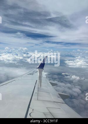 View from airplane window showing the wing above clouds under a partly cloudy blue sky. Stock Photo