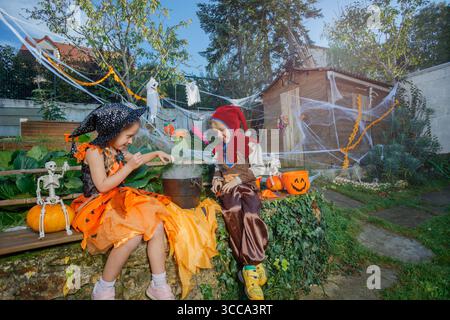 Two kids in Halloween costumes sit near steaming black cauldron Stock Photo