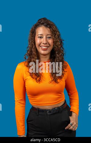 Studio portrait of a cheerful young latin woman with curly hair, wearing an orange shirt and black pants, posing confidently against a vibrant blue ba Stock Photo