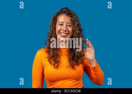 Studio portrait of a cheerful young woman with curly hair, wearing an orange shirt, waving and smiling against a vibrant blue backdrop Stock Photo