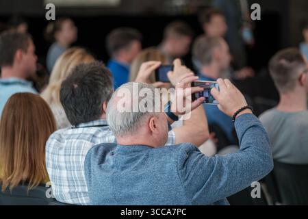 Audience sitting at an event with some attendees using smartphones to capture moments. Individuals focusing on the ongoing activity, fostering interaction and interest. Stock Photo