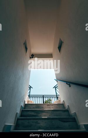 Low-angle view of marble stairs leading to an open sky, framed by white walls and black railings. Minimalist architectural design with natural light a Stock Photo