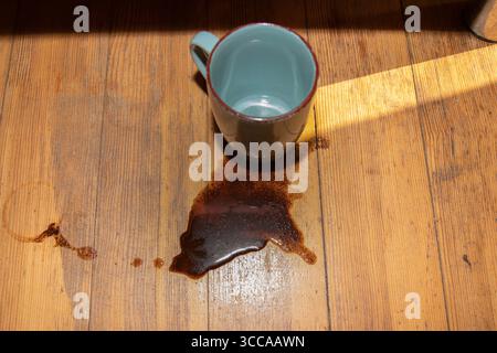 Close-up of spilled coffee from an empty turquoise ceramic mug on a rustic wooden floor in sunlight. Concept of accident, mess, or morning mishap Stock Photo