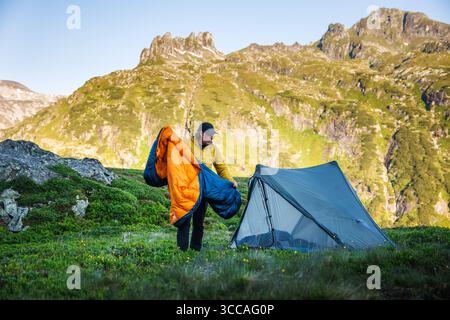 Camping in mountains. Man hiker putting sleeping bag into tent during trekking and mountain hiking Stock Photo