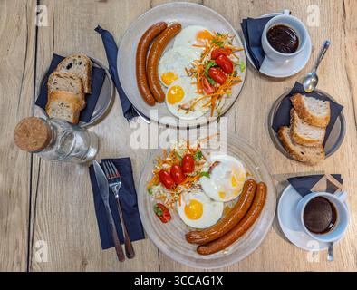Top-down view of a hearty breakfast for two, with fried eggs, sausages, fresh salad, bread, and coffee on a wooden table.Full traditional breakfast fo Stock Photo
