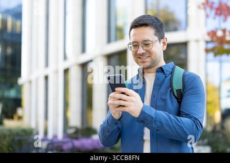 A cheerful individual interacts with a smartphone while standing in front of a contemporary building. The image conveys positivity, modern communication Stock Photo