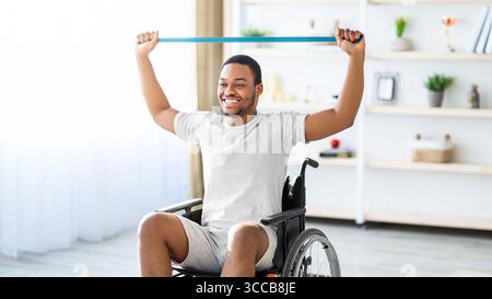 Happy black with disability man in wheelchair doing exercises with rubber band at home Stock Photo