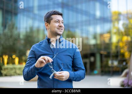 Man in a stylish blue shirt holding glasses and smiling with a glass building in the background. A positive and confident atmosphere in a modern urban setting. Stock Photo