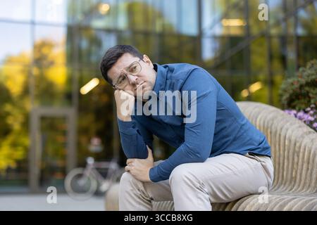 A person wearing casual clothing sits on a wooden bench outside, looking pensive. The background features a glass-paneled modern building, autumnal trees, and a parked bicycle. Stock Photo