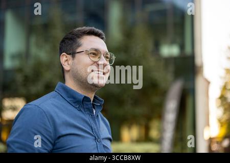 A cheerful man wearing glasses and a blue shirt standing outdoors near modern architecture. The image exudes positivity and professionalism, capturing an individual enjoying a moment in an urban Stock Photo